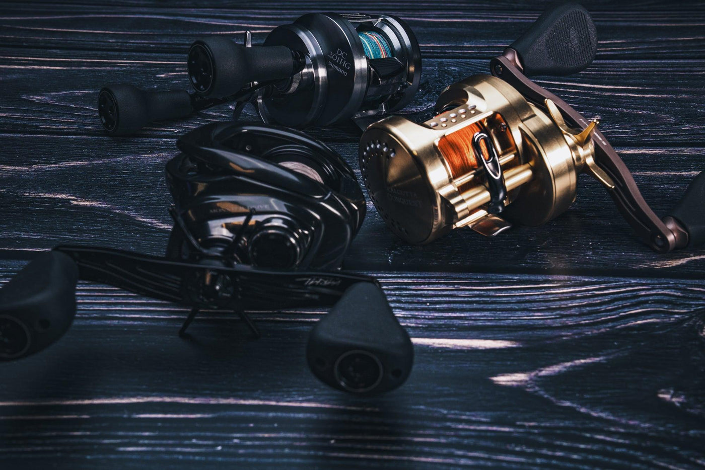 Drei Angelrollen, darunter eine glänzende goldene Rolle und zwei schwarze Rollen, sind auf einer dunklen, strukturierten Holzoberfläche platziert. Die Rollen sind dicht nebeneinander angeordnet und weisen unterschiedliche Designs und Griffe auf.