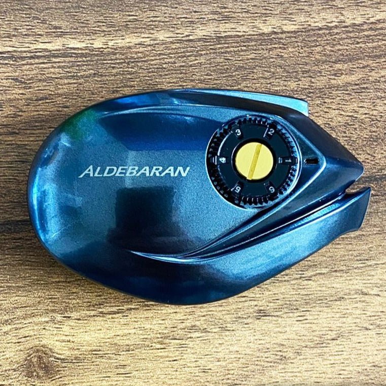 Eine Nahaufnahme der Hedgehog Studio Brake Dial Screw für Shimano 22 Aldebaran BFS in Gold und Schwarz, die für die Feinabstimmung der Bremseinstellung konzipiert und aus leichtem Duraluminium gefertigt ist und auf einer Holzoberfläche ruht.