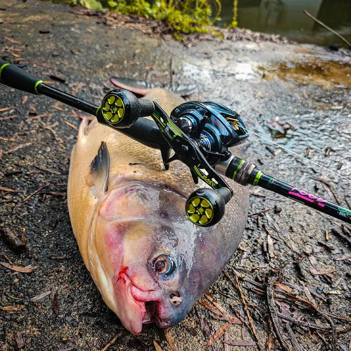 Ein großer Fisch liegt auf dem feuchten Boden, eine Angelrute und eine Rolle mit einem Gomexus Camo Carbon Handle (95 mm) und 27 mm TPE-Knöpfen (8 x 5 mm) liegen quer über seinem Körper. Zweige und Blätter liegen verstreut in der Nähe und dahinter schimmert Wasser.