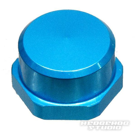 Eine metallisch blaue, sechseckige Duraluminium-Griffmutterkappe (Hedgehog Studio Shimano Handle Locknut M7 | für Original Shimano Kurbeln) mit glatter, abgerundeter Oberseite ist schräg vor einem weißen Hintergrund abgebildet. Unten rechts ist schwach „HEDGEHOG STUDIO“ zu sehen.