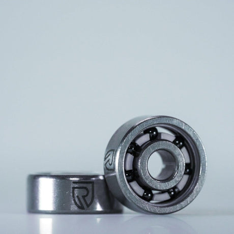 Zwei metallische RL-TNG High Speed Keramik Kugellager von RL-Angelrollentuning, gekennzeichnet mit dem stilisierten R-Logo, sind auf einem hellen Hintergrund abgebildet – eines aufrecht und gibt den Blick auf die Kugeln frei, eines flach dahinter. ABEC 9, alle Größen, für Baitcaster-Angelrollen.