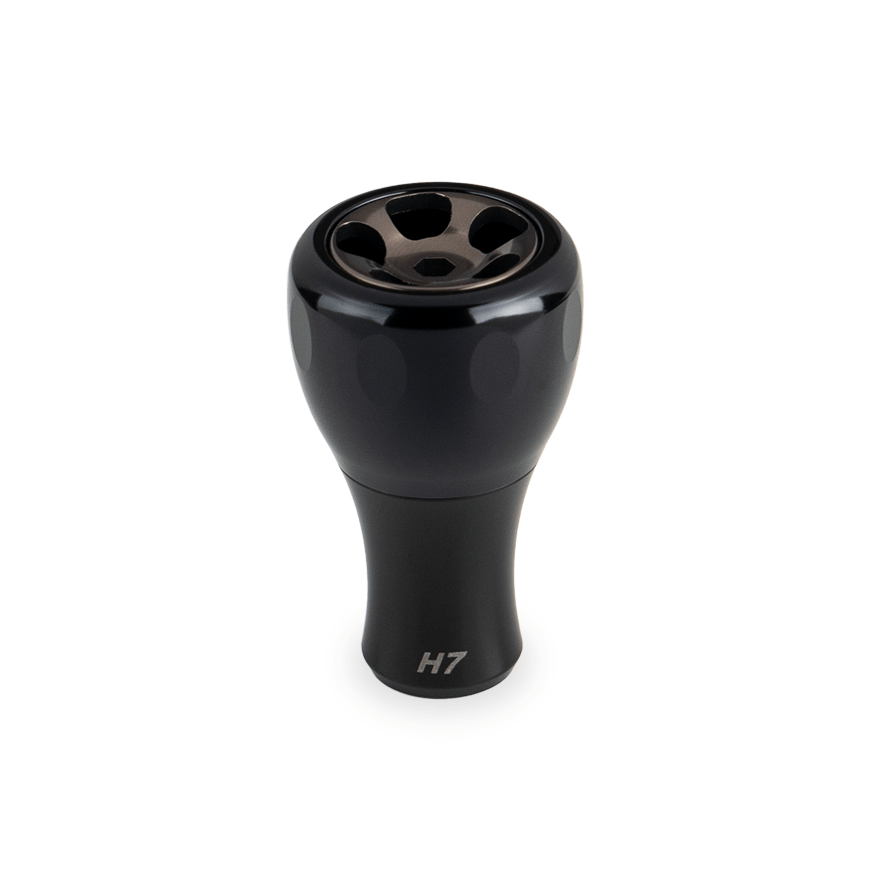 Ein schwarzer Taken H7 Knob 40mm - Aluminium Tuning Knauf, entworfen für Shimano & Daiwa Baitcaster und Spinnrollen, wird vor einem weißen Hintergrund gezeigt. Die metallisch glänzende Oberseite weist fünf kreisförmige Öffnungen und ein Branding im unteren Teil auf.