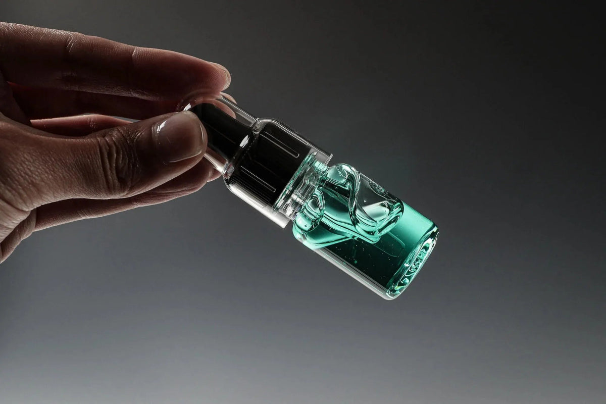 Eine Hand hält eine kleine Tropfflasche aus Glas mit Glitch Oil High Spec Bearing von Glitch Oil Japan, gefüllt mit transparenter blaugrüner Flüssigkeit und mit einer schwarzen Tropfkappe, vor einem dunklen Hintergrund mit Farbverlauf, von der Seite gezeigt.