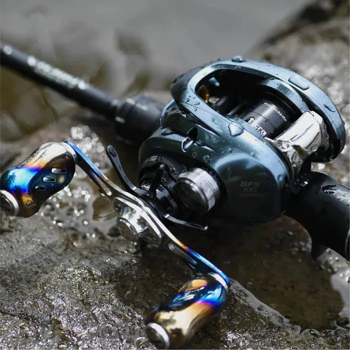 Eine Nahaufnahme der Gomexus-Aluminiumspule in einer Shimano 22 Aldebaran BFS-Rolle. Ihre Griffe und Spule weisen ein auffälliges metallisches, mehrfarbiges Finish mit Wassertropfen auf der Oberfläche auf, während sie auf einem nassen Felsen am Wasser ruht.