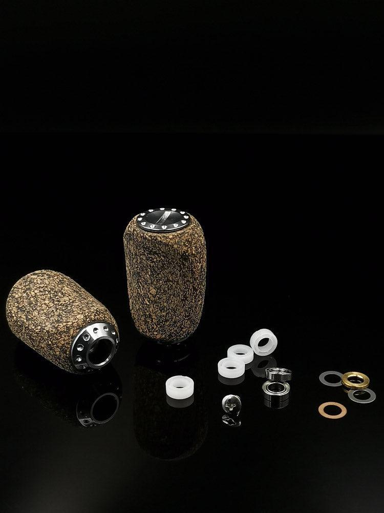 Zwei RL-Angelrollentuning RL-TNG Kork Knob 20mm | Slim | Universal Fit Knöpfe mit metallischen Enden ruhen auf einer schwarzen Oberfläche, umgeben von weißen Kunststoffringen, Edelstahlkugellagern, Metallscheiben und kreisförmigen Metallteilen.