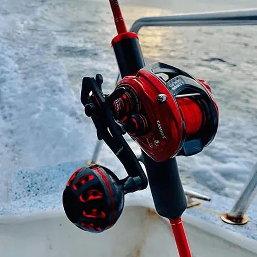 Eine Nahaufnahme einer roten Angelrolle mit dem Gomexus Carbon Handle 75 mm mit 38 mm Power Knob (8x5mm), die an einer Rute auf einem Boot befestigt ist. Im Hintergrund plätschert das Wasser, und eine Metallreling ist zu sehen.