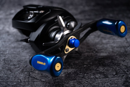 Nahaufnahme der RL-TNG EGL 95 Carbon Handle Baitcaster-Kurbel von RL-TNG mit elegantem schwarz-blauem Design mit goldenen Akzenten, leichtem Carbongriff und blauen Griffen mit SD Works-Beschriftung auf einer strukturierten dunklen Oberfläche.