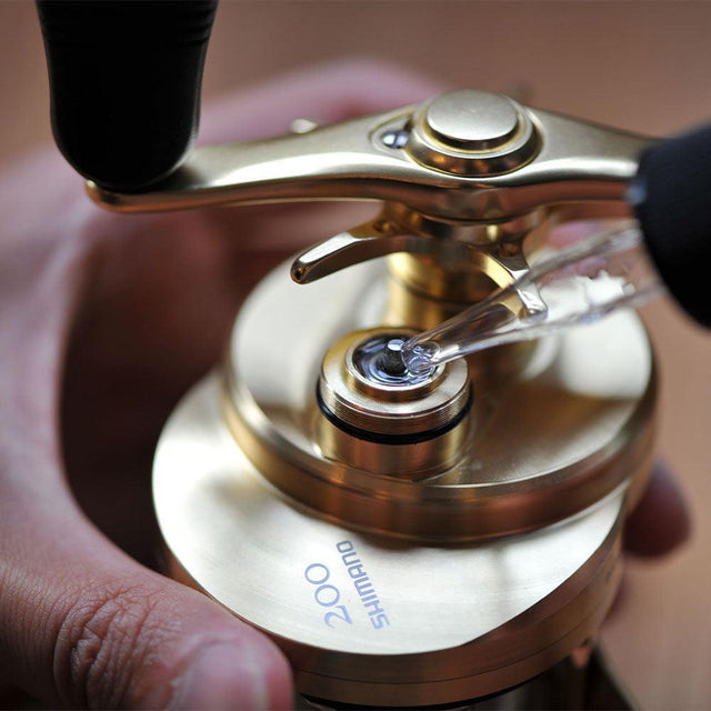 Eine Hand hält eine Shimano 200 Angelrolle, während mit einer Pipette RL-Angelrollentuning Premiumöl aus dem Baitcaster Wartung ohne Formular-Service für professionelle Rollenpflege und Ultraschallreinigung aufgetragen wird.