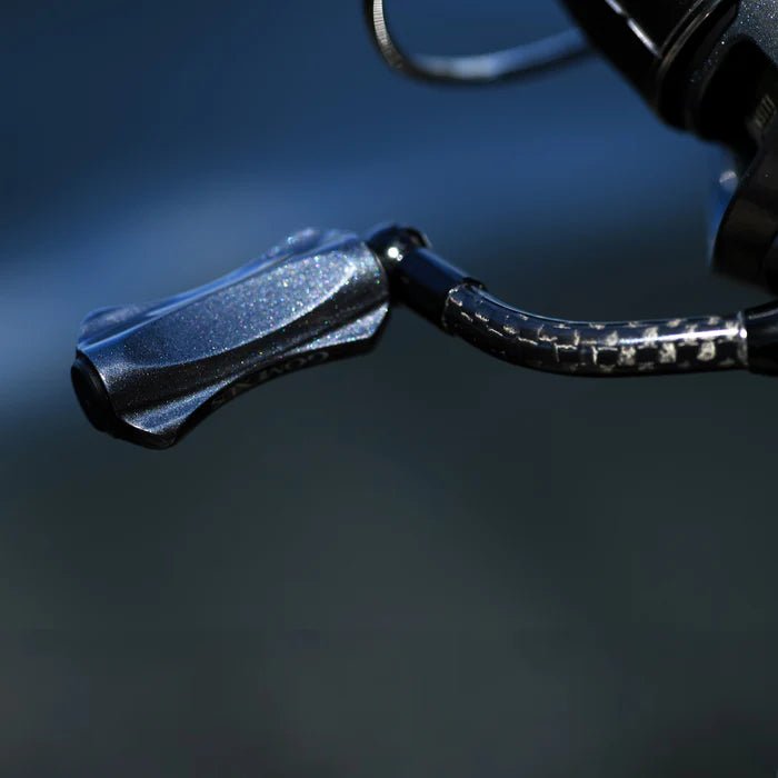 Eine Nahaufnahme des Gomexus CraftFin-35 Carbongriffs für Stationärrollen, mit einem 35mm Carbon-Kurbelarm und einem 18mm-Knauf für ultraleichte Angelrollen, angebracht an einer Rolle vor einem unscharfen dunklen Hintergrund.