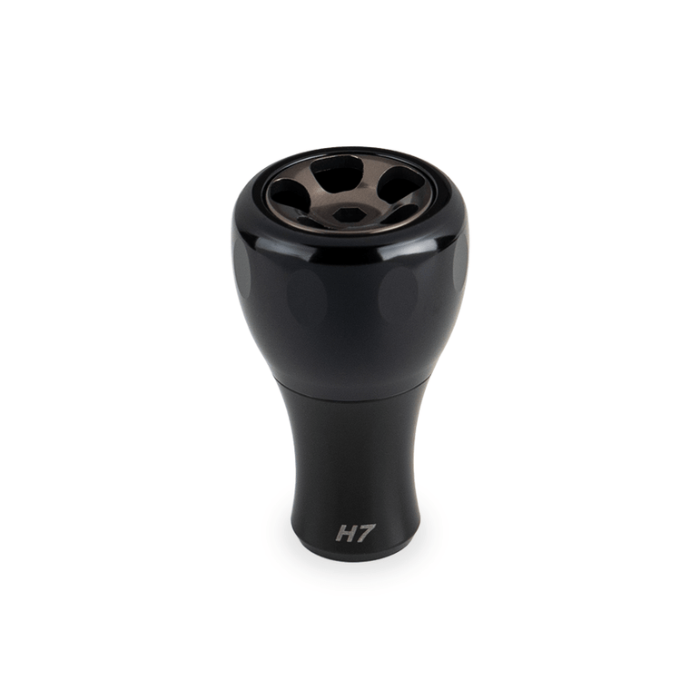 Ein schwarzer Taken H7 Knob 40mm - Aluminium Tuning Knauf, entworfen für Shimano & Daiwa Baitcaster und Spinnrollen, wird vor einem weißen Hintergrund gezeigt. Die metallisch glänzende Oberseite weist fünf kreisförmige Öffnungen und ein Branding im unteren Teil auf.