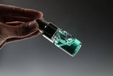 Eine Hand hält eine kleine Tropfflasche aus Glas mit Glitch Oil High Spec Bearing von Glitch Oil Japan, gefüllt mit transparenter blaugrüner Flüssigkeit und mit einer schwarzen Tropfkappe, vor einem dunklen Hintergrund mit Farbverlauf, von der Seite gezeigt.