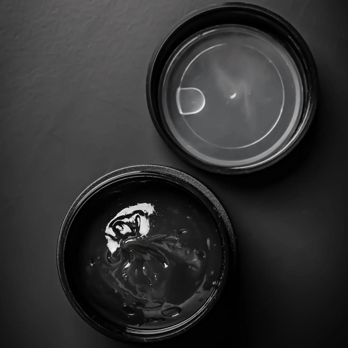 Ein offener schwarzer Behälter mit Glitch Oil Japans Premium Materia Grease Nr. 305, gefüllt mit glänzendem schwarzem Gel, steht auf einem dunklen Untergrund. Der umgedrehte Deckel weist innen Kondenswasser auf. Ideal für die Wartung von Angelrollen.