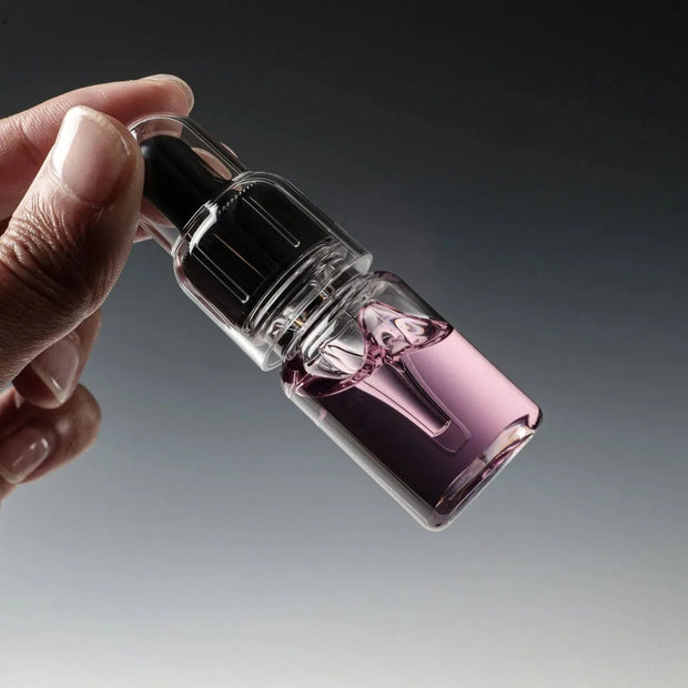 Eine Hand hält eine kleine Tropfflasche mit Glitch Oil Rosa von Glitch Oil Japan, gefüllt mit einer klaren, violett getönten Flüssigkeit, vor einem grauen Hintergrund mit Farbverlauf. Ideal zur Reduzierung von Korrosion und Verschleiß, insbesondere beim Salzwasserfischen.