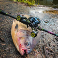 Ein großer Fisch liegt auf dem feuchten Boden, eine Angelrute und eine Rolle mit einem Gomexus Camo Carbon Handle (95 mm) und 27 mm TPE-Knöpfen (8 x 5 mm) liegen quer über seinem Körper. Zweige und Blätter liegen verstreut in der Nähe und dahinter schimmert Wasser.