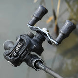 Eine Nahaufnahme einer schwarzen Angelrolle mit Wassertropfen, mit dem Gomexus Carbon Forged F24 Knob von Gomexus in ergonomischem Design, befestigt an einer über Wasser gehaltenen Rute mit verschwommenem Schilf im Hintergrund.