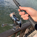 Eine Hand hält eine Angelrute mit einem Gomexus-Doppelgriff 98–82 mm mit T20-Knöpfen für Shimano, positioniert über dem Wasser neben einem Metallgeländer.