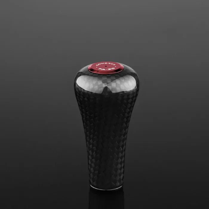 Ein Gomexus FA30 Carbon-Knopf aus Toray-Carbon mit strukturiertem Muster und roter runder Oberseite steht aufrecht auf einer reflektierenden schwarzen Oberfläche.