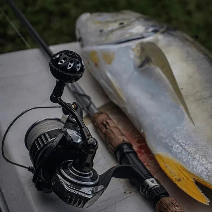 Eine Angelrute mit einem Gomexus Plug & Play Aluminium Power Handle für Daiwa BG MQ liegt auf einer Oberfläche neben einem großen, frisch gefangenen silber-gelben Fisch und deutet auf einen kürzlichen Fang in der Nähe des Wasserrands hin.