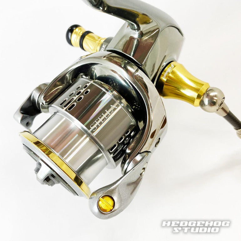 Nahaufnahme einer metallischen Hedgehog Studio Extra Super Duralumin Schraube für Schnurlaufröllchen mit goldenen und silbernen Akzenten vor einem schlichten weißen Hintergrund. Das Hedgehog Studio-Logo ist in der unteren rechten Ecke sichtbar.