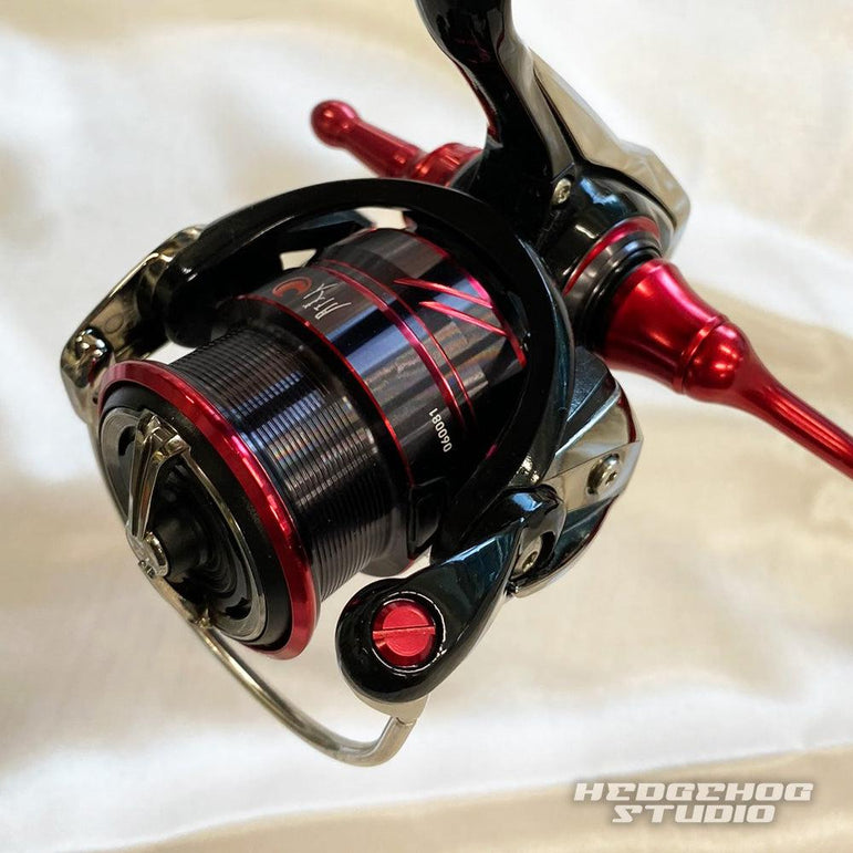 Eine Nahaufnahme einer schwarz-roten Angelrolle „Hedgehog Studio Extra Super Duralumin Schraube für Schnurlaufröllchen“ mit metallischen Akzenten auf einer hellen Oberfläche; die Spule und der Duraluminiumgriff sind sichtbar, mit dem Aufdruck „Hedgehog Studio“ unten rechts.