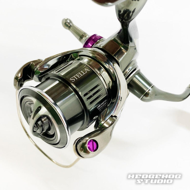 Ein Nahaufnahmefoto zeigt eine Shimano Stella-Rolle mit detaillierten Teilen, violetten Akzenten und einem Hedgehog Studio Extra Super Duralumin Schraube für Schnurlaufröllchen-Upgrade mit den Logos STELLA und HEDGEHOG STUDIO auf der Rolle.