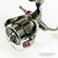 Ein Nahaufnahmefoto zeigt eine Shimano Stella-Rolle mit detaillierten Teilen, violetten Akzenten und einem Hedgehog Studio Extra Super Duralumin Schraube für Schnurlaufröllchen-Upgrade mit den Logos STELLA und HEDGEHOG STUDIO auf der Rolle.