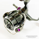 Ein Nahaufnahmefoto zeigt eine Shimano Stella-Rolle mit detaillierten Teilen, violetten Akzenten und einem Hedgehog Studio Extra Super Duralumin Schraube für Schnurlaufröllchen-Upgrade mit den Logos STELLA und HEDGEHOG STUDIO auf der Rolle.