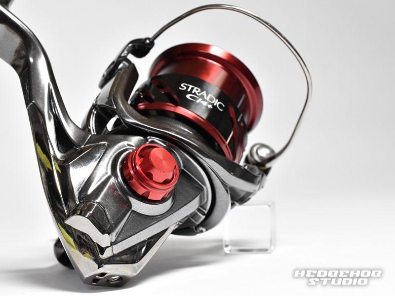 Nahaufnahme der Hedgehog Studio Handle Screw Cap von Hedgehog Studio auf einer Shimano Stradic CI4+ Spinnrolle mit metallisch-silbernen und roten Akzenten. Das Hedgehog Studio-Logo erscheint unten rechts auf weißem Untergrund.