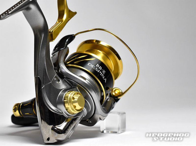 Eine Nahaufnahme einer gold-schwarzen Angelrolle mit einer Schraubkappe am Griff von Hedgehog Studio, die auf einer weißen Oberfläche neben einem kleinen durchsichtigen Block und dem Logo von Hedgehog Studio in der Ecke ruht.