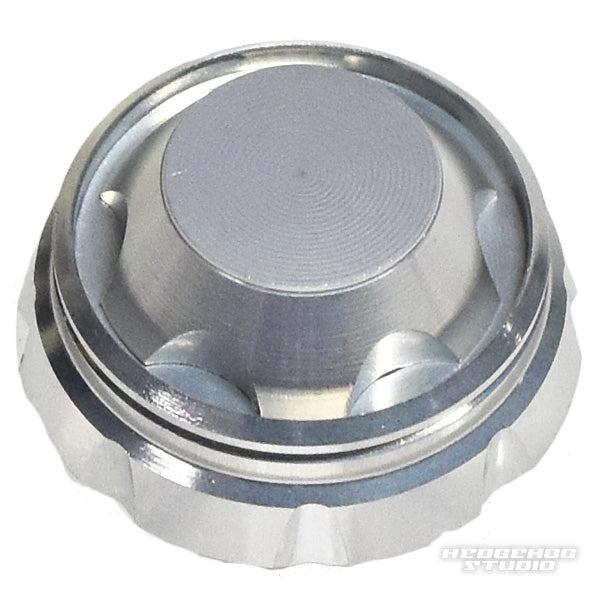 Eine Nahaufnahme des Schraubverschlusses von Hedgehog Studio mit rundem silbernem Design, strukturiertem Rand und flacher Oberseite auf weißem Untergrund. Das Hedgehog Studio-Logo befindet sich in der unteren rechten Ecke.