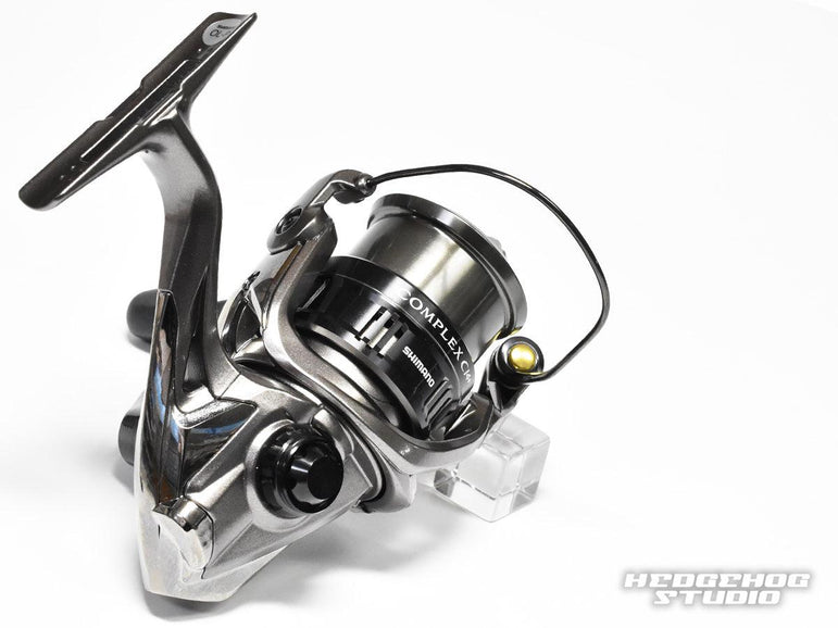 Nahaufnahme einer metallischen Angelrolle mit schwarzer Spule und Griff, ausgestattet mit dem Hedgehog Studio-Griffschraubverschluss. Die Rolle trägt das COMPLEX-Logo, während das Hedgehog Studio-Logo in der unteren rechten Ecke erscheint.