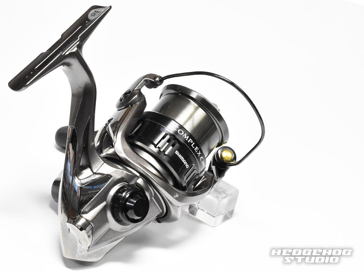 Nahaufnahme einer metallischen Angelrolle mit schwarzer Spule und Griff, ausgestattet mit dem Hedgehog Studio-Griffschraubverschluss. Die Rolle trägt das COMPLEX-Logo, während das Hedgehog Studio-Logo in der unteren rechten Ecke erscheint.