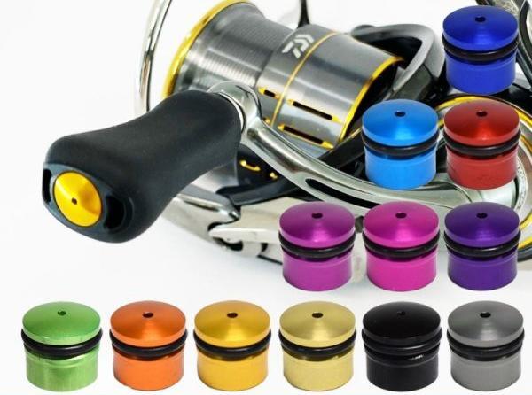 Ein Hedgehog Studio Knob Cap | Daiwa Finesse Knob ist neben einer Angelrolle mit schwarzem Griff abgebildet, umgeben von zwölf metallisch gefärbten Duraluminiumknöpfen in Blau, Lila, Rot, Grün, Gold, Silber und Schwarz.