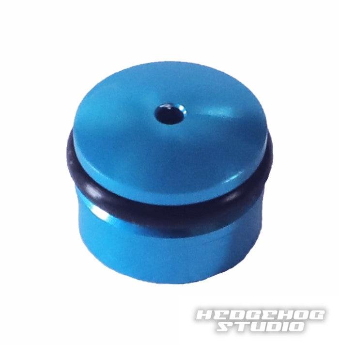 Eine kleine, runde, blaue Hedgehog Studio Knob Cap für den Daiwa Finesse Knob mit zentralem Loch und schwarzem O-Ring ist auf weißem Hintergrund abgebildet. Das Hedgehog Studio Logo erscheint in der unteren rechten Ecke.