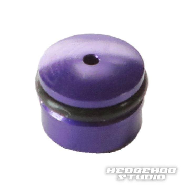 Eine kleine, zylindrische, metallisch-lila Hedgehog Studio-Knopfkappe für Daiwa Finesse-Knöpfe mit einem zentralen Loch oben und einem schwarzen O-Ring ist auf einem weißen Hintergrund mit dem Hedgehog Studio-Logo in der unteren rechten Ecke abgebildet.