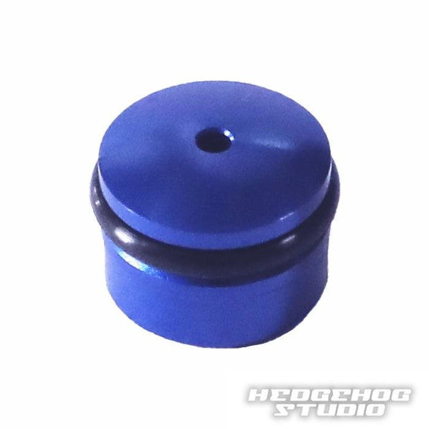 Eine kleine blaue zylindrische Hedgehog Studio-Knopfkappe für Daiwa Finesse-Knöpfe aus Duralumin mit einem zentralen Loch und einem schwarzen O-Ring wird auf einem weißen Hintergrund mit dem Hedgehog Studio-Logo in der Ecke angezeigt.