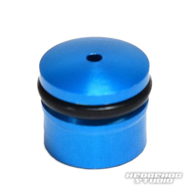 Eine kleine, zylindrische blaue Hedgehog Studio-Knopfkappe für Daiwa Zaion aus Duralumin mit abgerundeter Oberseite und einem schwarzen O-Ring in der Mitte ist auf einem weißen Hintergrund mit dem Hedgehog Studio-Logo in der unteren rechten Ecke abgebildet.
