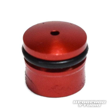 Eine kleine, zylindrische, rote Hedgehog Studio Knob Cap (Daiwa Zaion) mit einem schwarzen Gummi-O-Ring im oberen Bereich und einem zentralen Loch auf der flachen Oberseite. Das Foto zeigt unten rechts ein Wasserzeichen von Hedgehog Studio.