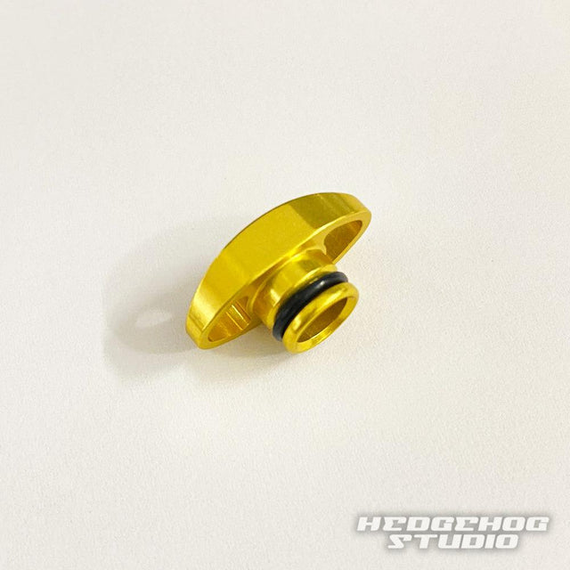 Eine kleine goldene Hedgehog Studio Knob Cap mit schwarzem O-Ring für Shimano 20 Metanium, 22 Metanium Shallow Edition und 20 SLX DC liegt auf einer weißen Fläche, unten rechts befindet sich das HEDGEHOG STUDIO Logo.