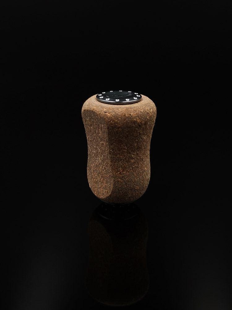 Der RL-TNG Kork Knob 20mm von RL-Angelrollentuning – ein einzelner Korkgriff mit schwarzer Folienoberfläche – steht aufrecht vor einem schwarzen Hintergrund und erzeugt eine subtile Reflexion darunter. Universelle Passform für vielseitigen Einsatz.