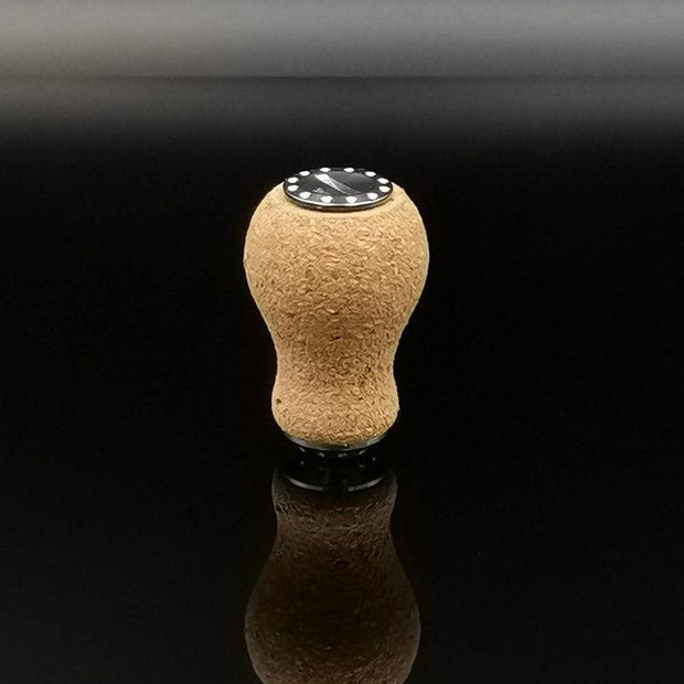 Der RL-TNG Kork Knob 20mm | Erdnuss | Universal Fit von RL-Angelrollentuning steht aufrecht auf einer reflektierenden schwarzen Oberfläche, sein abgerundeter Korkstopfen und die schwarze runde Basis spiegeln sich darunter.