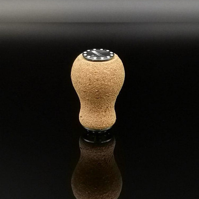 Der RL-TNG Kork Knob 20mm | Erdnuss | Universal Fit von RL-Angelrollentuning steht aufrecht auf einer reflektierenden schwarzen Oberfläche, sein abgerundeter Korkstopfen und die schwarze runde Basis spiegeln sich darunter.