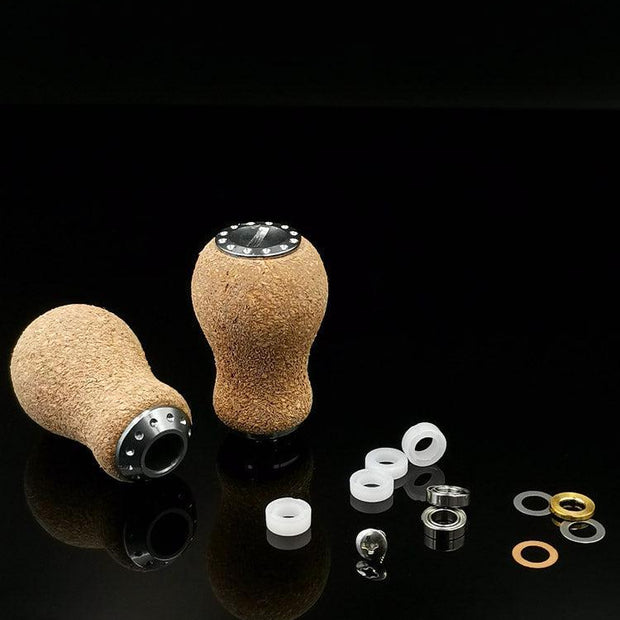 Zwei RL-TNG Kork Knob 20mm Peanut Schaltknäufe von RL-Angelrollentuning mit Metallakzenten werden mit Unterlegscheiben, Muttern und Ringen gezeigt – ideal zum Individualisieren Ihrer Angelrolle oder Ihres Edelstahlkugellagers. Präsentiert auf einer glänzend schwarzen Oberfläche.