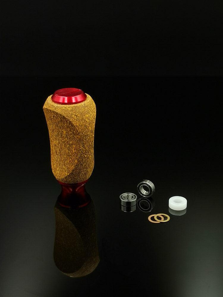 Der RL-TNG Kork Knob von RL-Angelrollentuning, ein mehrfarbiger Universalgriff aus Holz und rotem Metall, steht aufrecht auf einer reflektierenden schwarzen Oberfläche neben kleinen Metalllagern, einem weißen Kunststoffteil und zwei dünnen kreisförmigen Ringen.