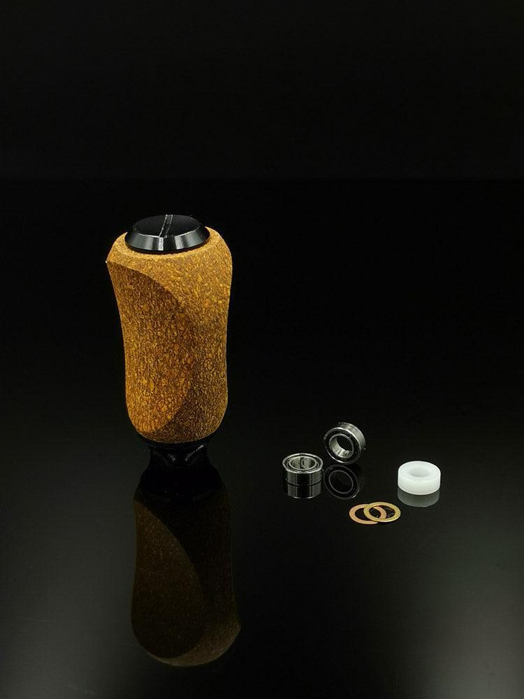 Ein RL-TNG Kork Knob von RL-Angelrollentuning mit einem mehrfarbigen braun-schwarzen Griff und universeller Passform steht auf einer reflektierenden Oberfläche. In der Nähe befinden sich einzelne Angelrollenteile: Lager, Unterlegscheiben, Metallring und weißer Zylinder.