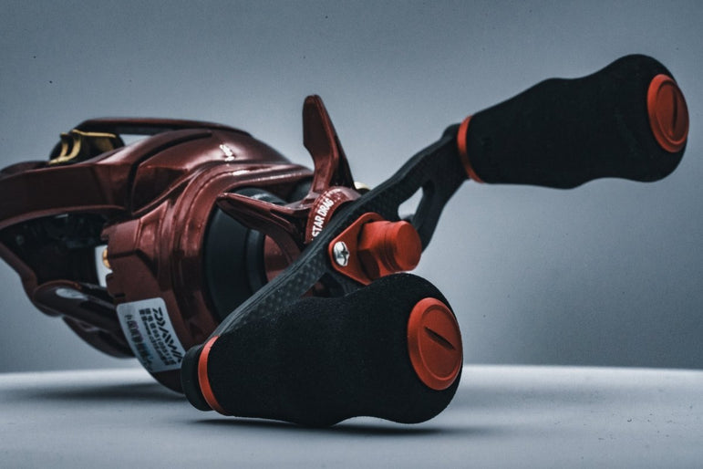 Eine Nahaufnahme der Angelrolle RL-TNG OCT 24 EVA Knob von RL-TNG mit rot-schwarzem Design, strukturierten schwarzen EVA-Knob-Griffen und orangefarbenen Akzenten auf einer schlichten Oberfläche mit grauem Hintergrund.