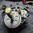 Nahaufnahme von mechanischen Zahnrädern und Teilen im Inneren einer teilweise demontierten Daiwa Tatula-Rolle, wobei ein Rollenwerk Dragclicker für Daiwa Tatula 70/80/100/103/SLP Works 105 zwischen verstreuten Komponenten auf einer grau strukturierten Oberfläche sichtbar ist.