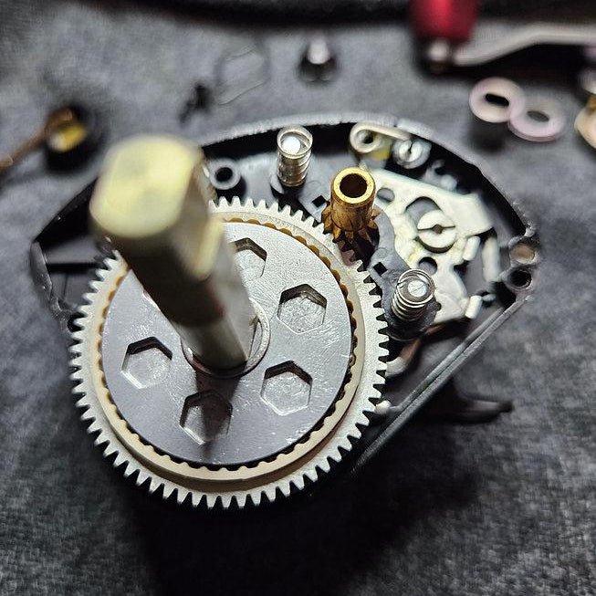 Nahaufnahme des Rollenwerk Dragclickers für Shimano SLX | Curado 71/151, wobei die Innenverzahnung, die Schrauben und die Metallwelle auf einer grauen, strukturierten Oberfläche hervorgehoben werden.