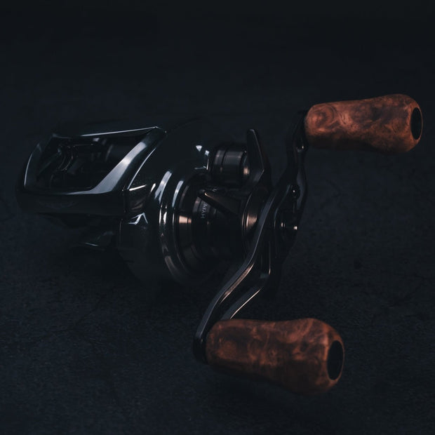 Nahaufnahme einer dunklen, schicken Baitcaster-Rolle mit den legendären SLP Works i Shape Wood Knob Limited Edition Griffen – edles Holz trifft cooles Metall! Das Ganze auf einem stylisch-texturierten Hintergrund, Licht bringt die feinen Details der Holzgriffe und die sexy Kurven Ihrer Tuning-Rolle perfekt zur Geltung. Perfekt für alle, die aus ihrer Angelrolle das Maximum rausholen wollen!.