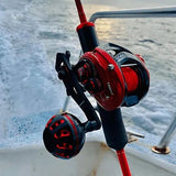 Eine Nahaufnahme einer roten Angelrolle mit dem Gomexus Carbon Handle 75 mm mit 38 mm Power Knob (8x5mm), die an einer Rute auf einem Boot befestigt ist. Im Hintergrund plätschert das Wasser, und eine Metallreling ist zu sehen.